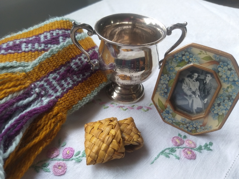 Several items arranged on a cloth embroidered with flowers: a yellow and purple knitted scarf, a silver-coloured trophy cup, a small bag woven from flax, and a black-and-white photograph of a seated couple in a frame decorated with blue flowers.