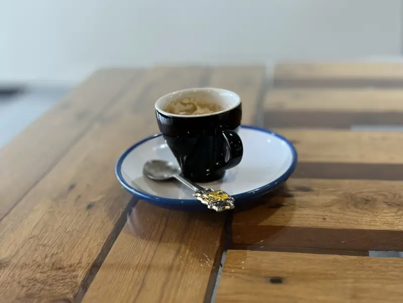 A black espresso cup with teaspoon on a white saucer, placed on a wooden table