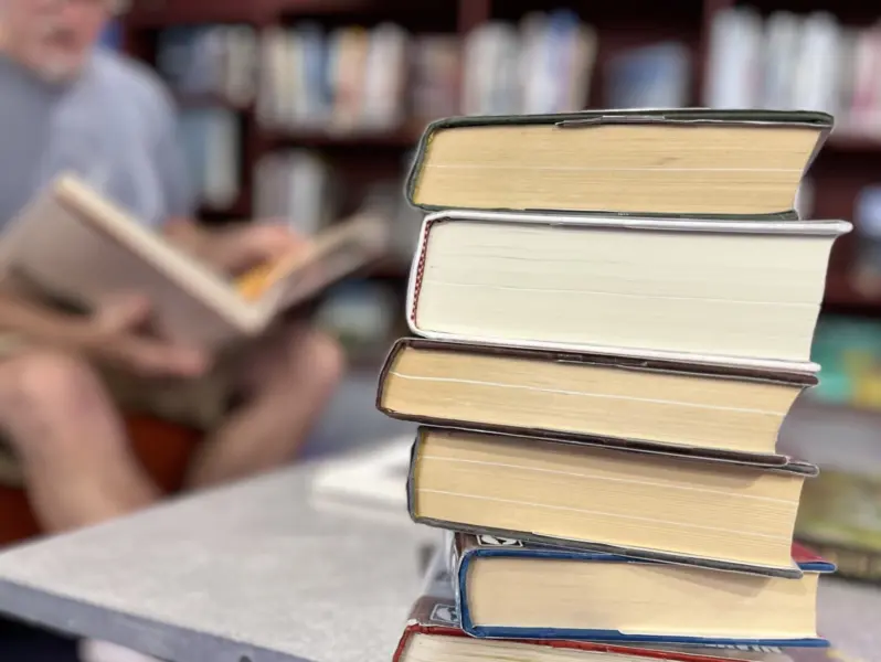 A stack of hardback books, spines facing away, with bookshelves and a figure reading in the background